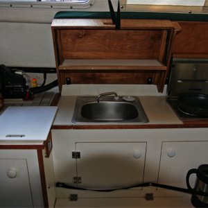 Covers are raised and helm seat pivots up to use galley. Hot items can be placed on ceramic tiles under helm seat. Small drop-down shelf inside forward cover is made of starboard trimmed in teak. Snap on elastic bands are handy for holding essentials (spelled COFFEE press) when underway.