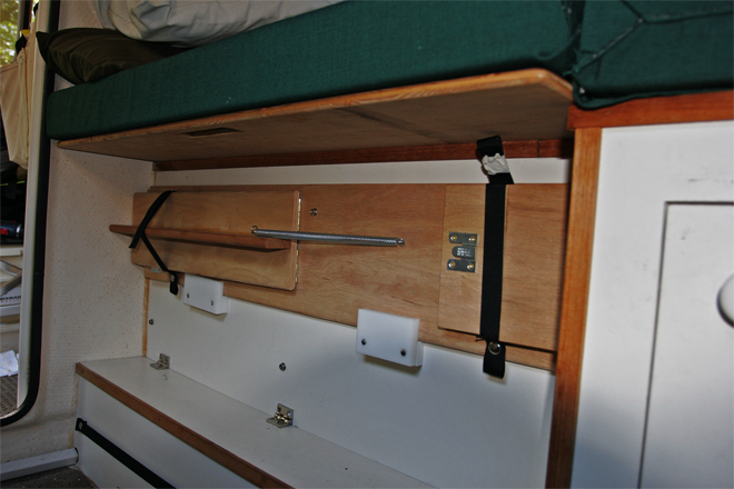 Bed support for the starboard bed is stowed on the  port-side box. Note the extender support  holding the port extender cushion. It can be quickly slid back under the lid to allow use of the original cushion for the 2 position commuter seat. The starboard bed support rests on a wood block at each end and is captured by the slotted fingers; it is rock solid with the leg extended. The storage box with lid is 40 inches long and holds kitchen items.